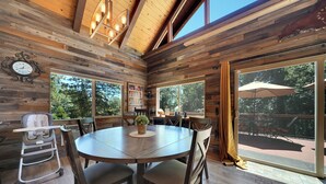 A wooden interior with a round table, chairs, a bookshelf, and a large window with an umbrella outside. - Tranquil Hillside Home w/ Private Hot Tub! (Pioneer)