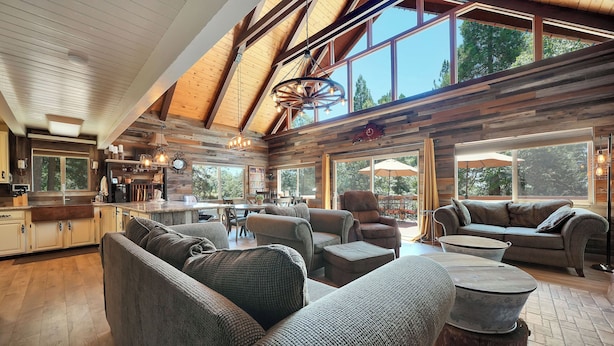 A spacious living area with a high ceiling, wooden beams, and large windows offering a view of the outdoors. - Tranquil Hillside Home w/ Private Hot Tub! (Pioneer)
