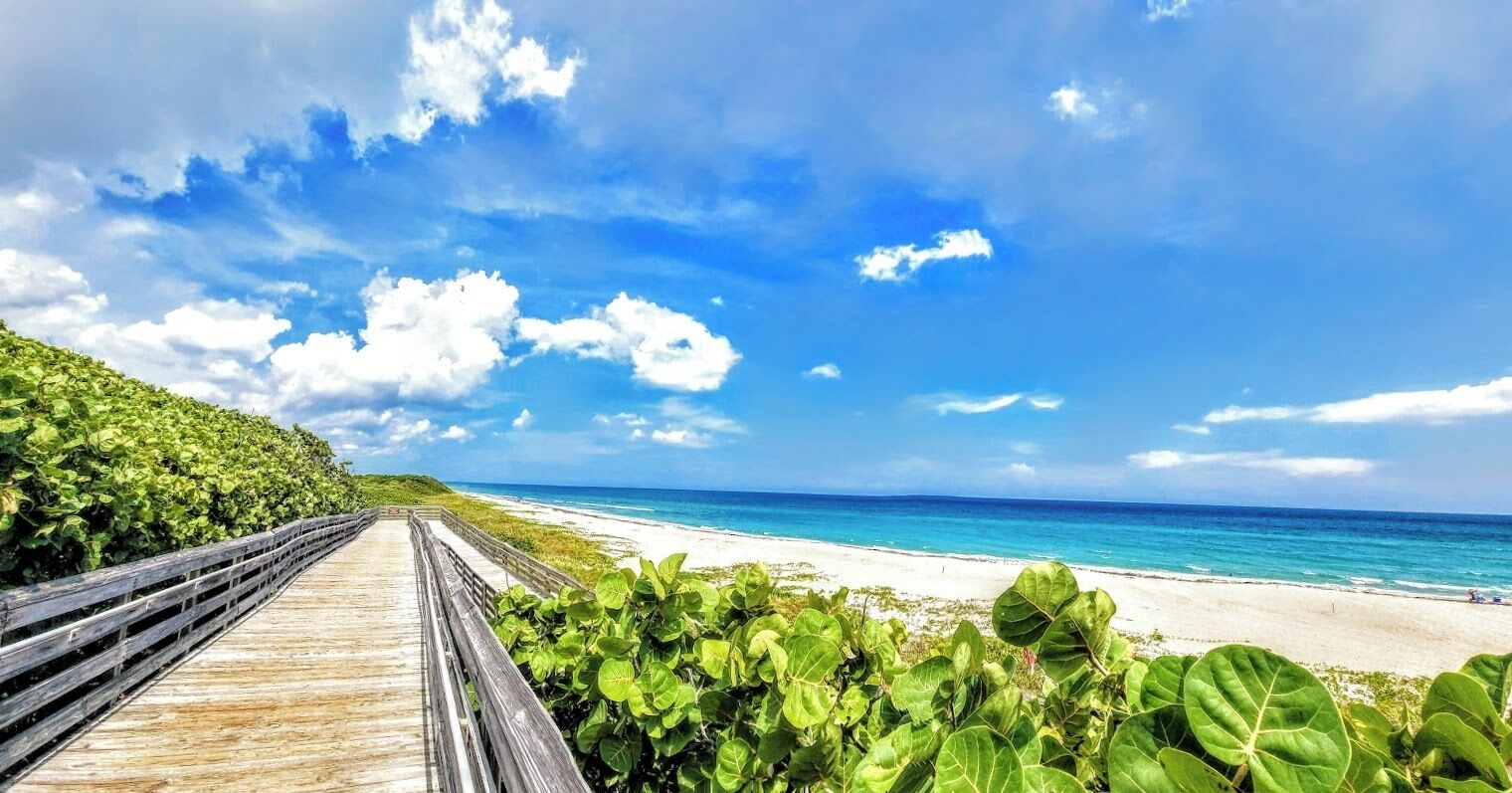 Une promenade en bois qui mène à une plage où l’eau est d’un bleu limpide et le ciel est partiellement nuageux.