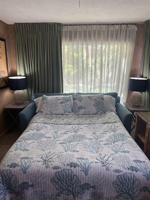 Ein Schlafzimmer mit einem Bett, zwei Nachttischen, einem Fenster mit leichtem Vorhang und Blick auf Grünfläche.