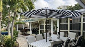 Un patio al aire libre con una mesa y sillas blancas, un paraguas a rayas y vista de un edificio al fondo.