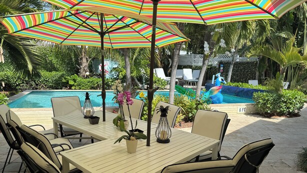 Zona junto a la piscina con mesa y sillas, una sombrilla de rayas coloridas y una piscina al fondo.