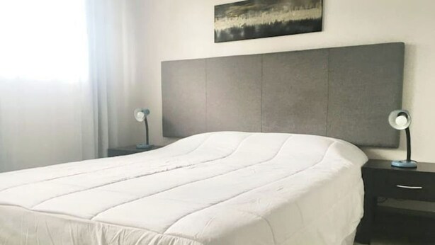 Ein ordentlich bezogenes Bett mit grauem Kopfteil, zwei Nachttische mit Lampen und ein Bild an der Wand.