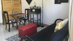 Ein kompakter Wohnbereich mit einer Couch, einem kleinen Tisch, zwei Stühlen, einem roten Hocker, einer Stehlampe und einem an der Wand montierten Regal mit Dekoration.