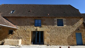 Um edifício de pedra com telhado em tijolos, uma entrada central com porta e duas janelas com persianas azuis.
