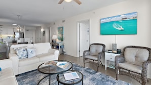 A modern living room with a white sofa, two wicker chairs, a coffee table, and a flat-screen TV displaying a beach scene.