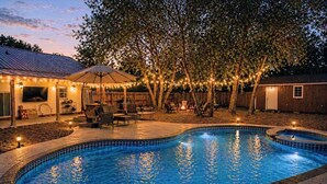 Un patio trasero con piscina, mobiliario de exterior e iluminación con luces colgantes.