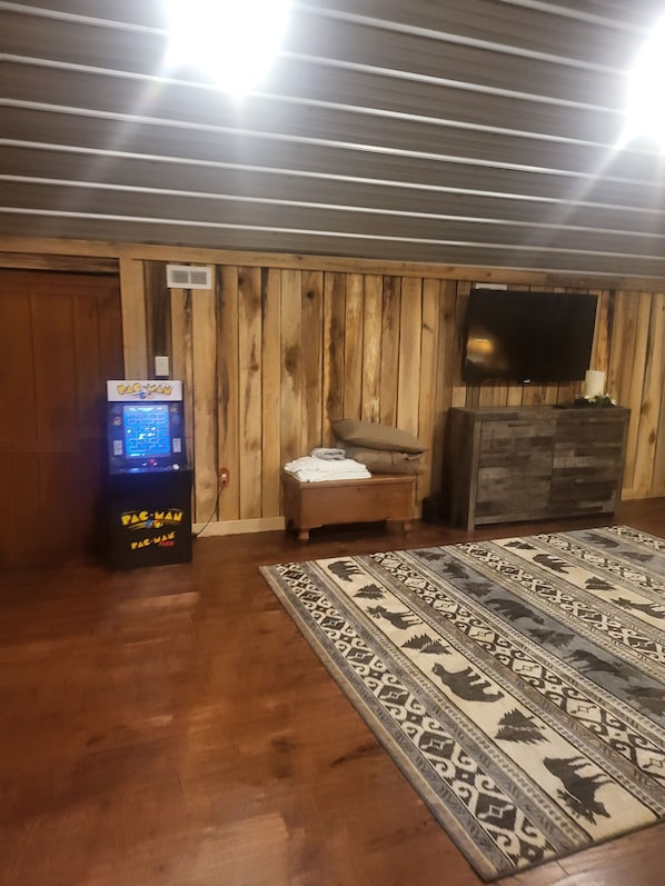 Une pièce aux murs recouverts de lambris en bois, un téléviseur à écran plat fixé au mur, une borne d’arcade Pac-Man, un petit canapé et un tapis à motifs.