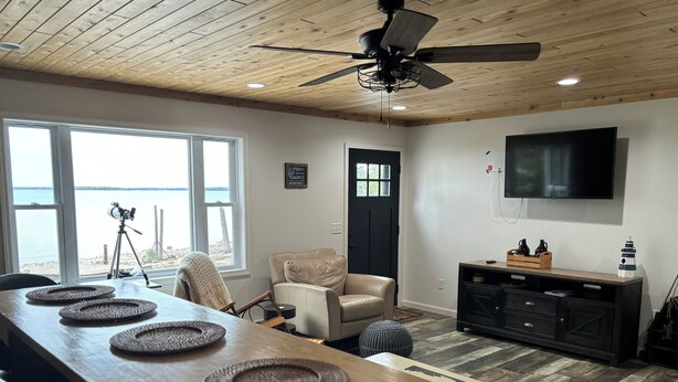 A room with a wooden ceiling, a ceiling fan, a large window with a view of the beach, a black door, a TV mounted on the wall, a wooden table with woven placemats, a leather chair, and a black cabinet with decorative items. - Beautiful, waterfront home located on the St Mary’s River, fisherman’s dream  (De Tour Village)