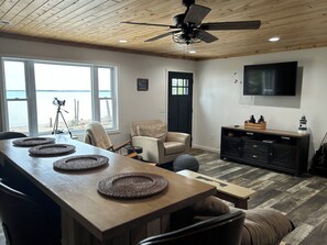 A room with a wooden ceiling, a ceiling fan, a large window with a view of the beach, a black door, a TV mounted on the wall, a wooden table with woven placemats, a leather chair, and a black cabinet with decorative items.