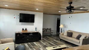 A modern living room with a wooden ceiling, a flat-screen TV mounted on the wall, a black entertainment unit, a beige sofa, and a wooden coffee table. - Beautiful, waterfront home located on the St Mary’s River, fisherman’s dream  (De Tour Village)