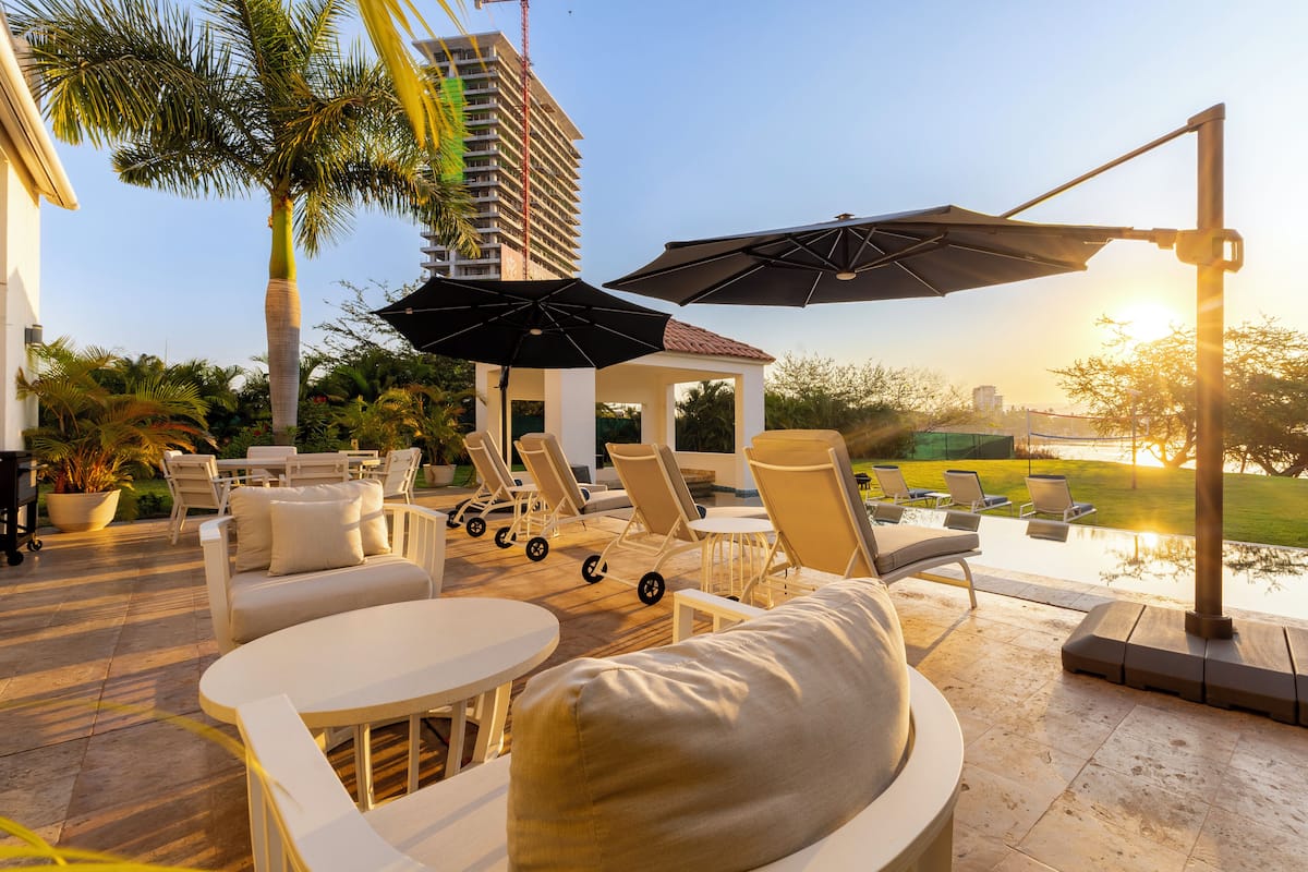 A sunlit outdoor lounge area with white furniture, umbrellas, and a view of a building and a body of water.