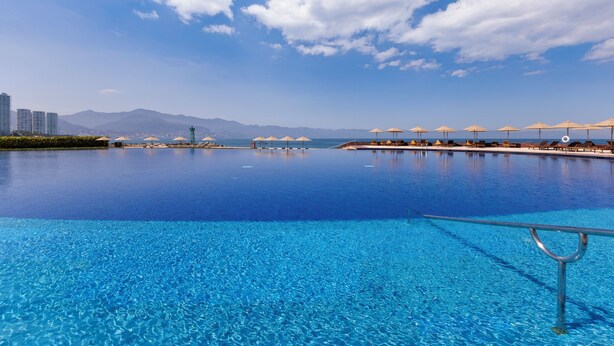 A large infinity pool with clear blue water, surrounded by beach umbrellas and lounge chairs, with mountains in the background. - Capitalia Tres Mares Beach Residences & Marina (Puerto Vallarta)