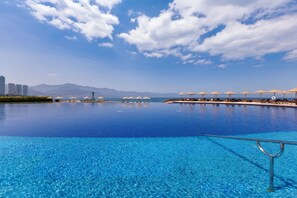 Une grande piscine à débordement à l’eau d’un bleu cristallin, entourée de parasols de plage et de chaises longues, avec des montagnes en toile de fond.