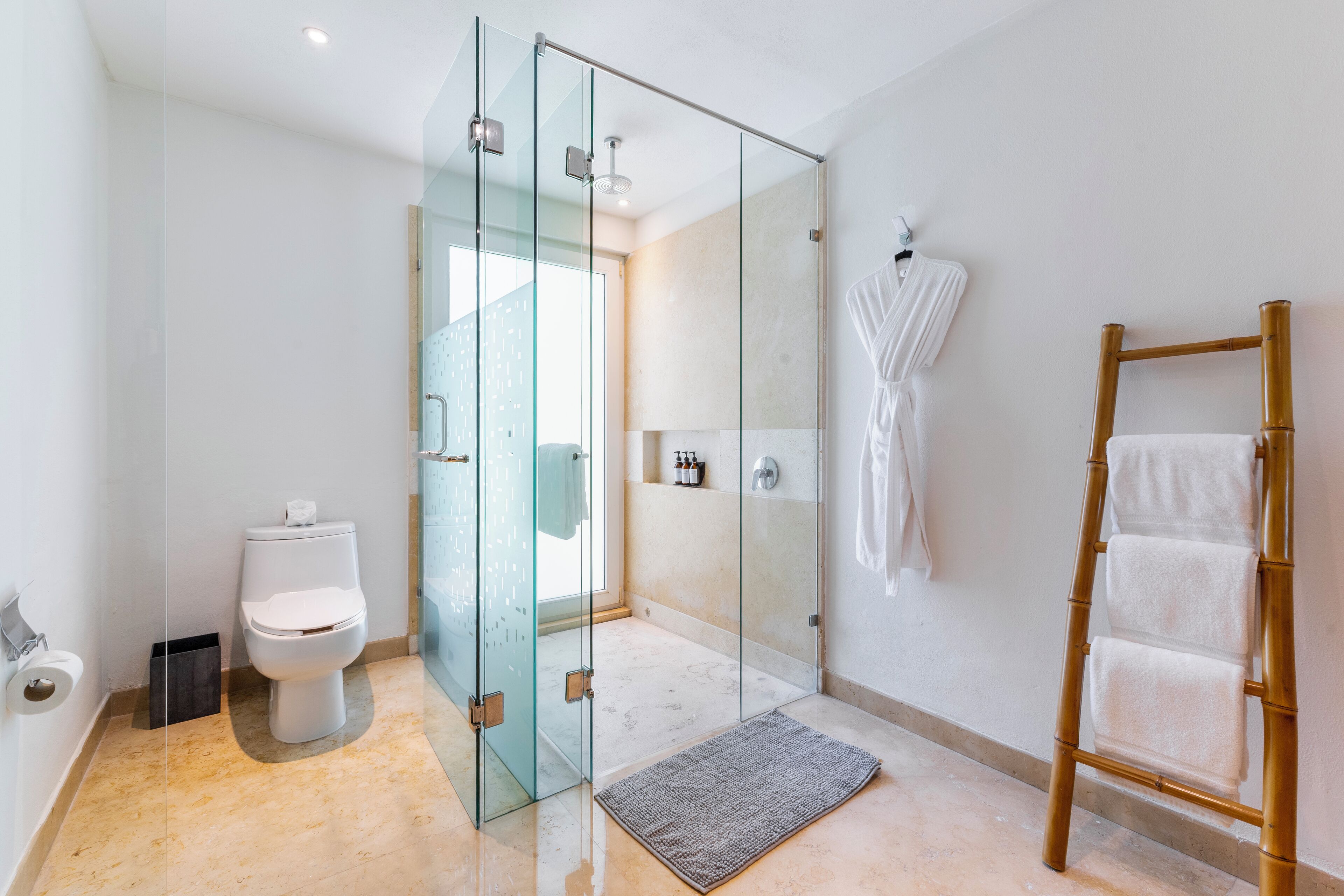 A modern bathroom with a glass shower enclosure, a toilet, a wooden ladder with towels, and a white robe hanging on the wall.