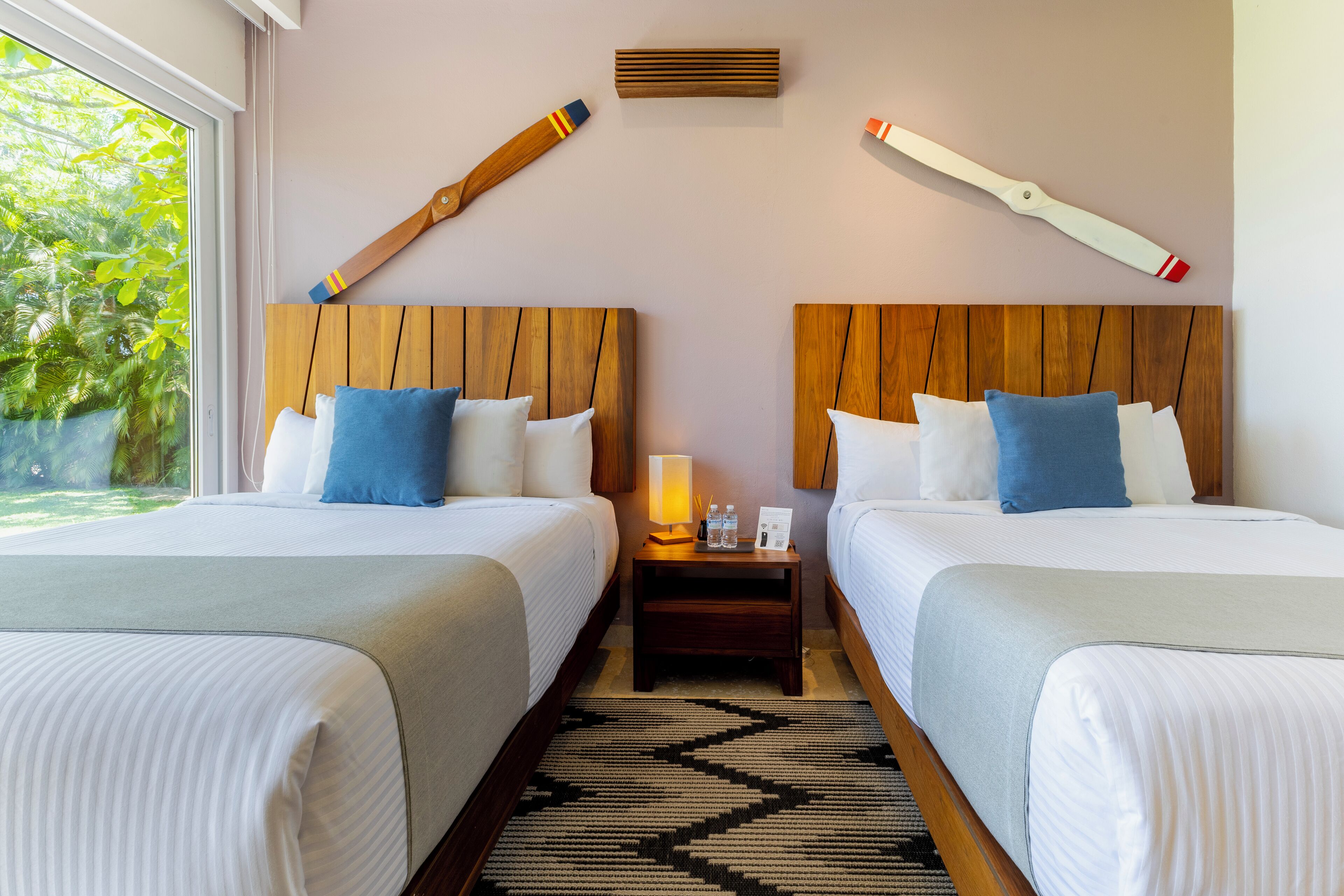 A hotel room with two beds, wooden headboards, a nightstand with a lamp, and a view of greenery outside.