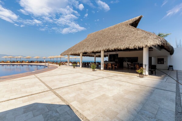 A poolside area with a thatched-roof structure, lounge chairs, and a view of the sea. - Capitalia Tres Mares Beach Residences & Marina (Puerto Vallarta)