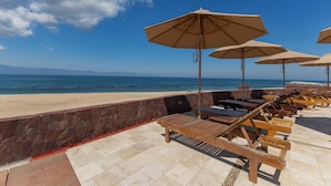 A beachside lounge area with wooden loungers and beige umbrellas, overlooking the sea. - Capitalia Tres Mares Beach Residences & Marina (Puerto Vallarta)