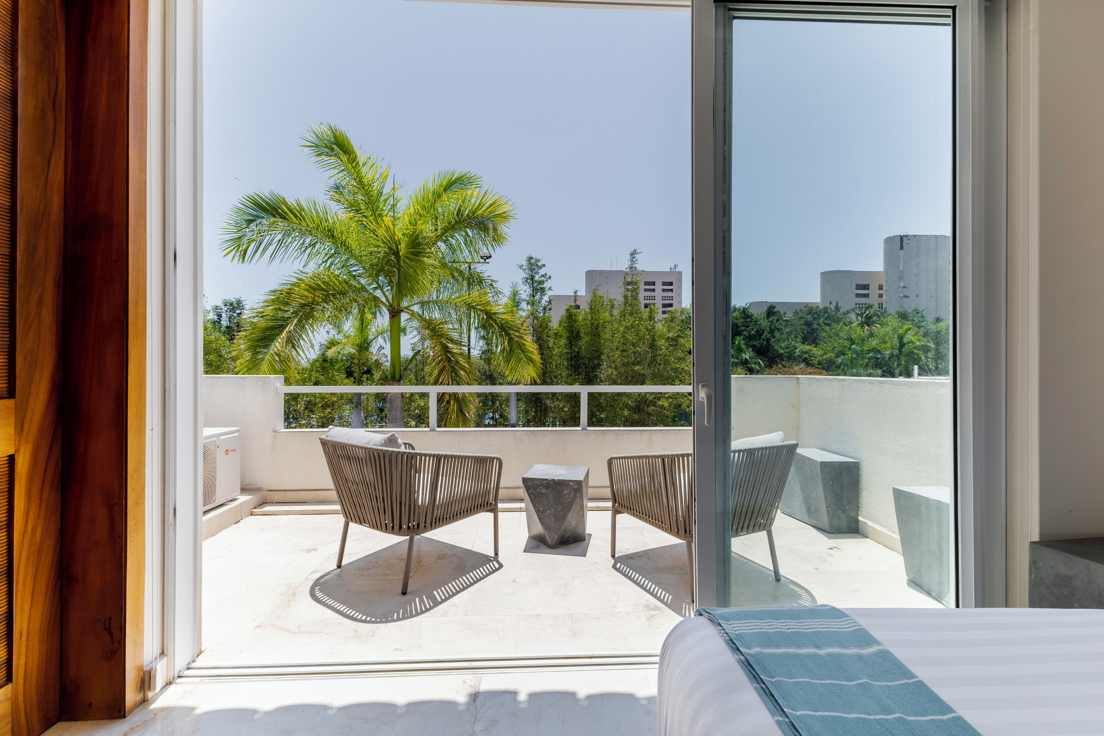 Un balcon équipé d’un lit, de deux chaises, d’une petite table et offrant une vue sur des palmiers et des immeubles.