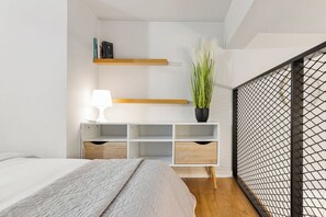 Quarto com cama, mesa de cabeceira com luminária, planta e estante com livros.