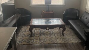 Un salón con piso de madera, una mesa de centro de vidrio, dos sofás de cuero negro, un televisor sobre un mueble bajo y una ventana con persianas.