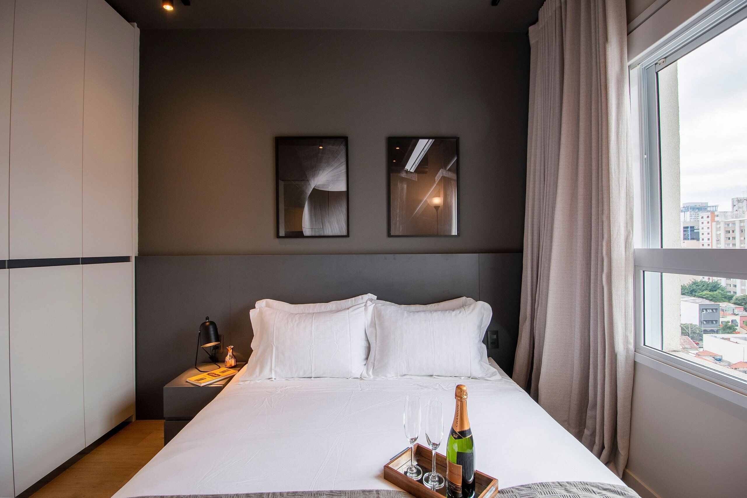 A hotel room with a large bed, two framed pictures on the wall, a nightstand with a lamp and a bottle, and a window with curtains.
