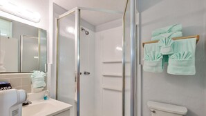 A bathroom with a glass shower enclosure, a toilet, a sink with a makeup mirror, and towels hanging on a rack.