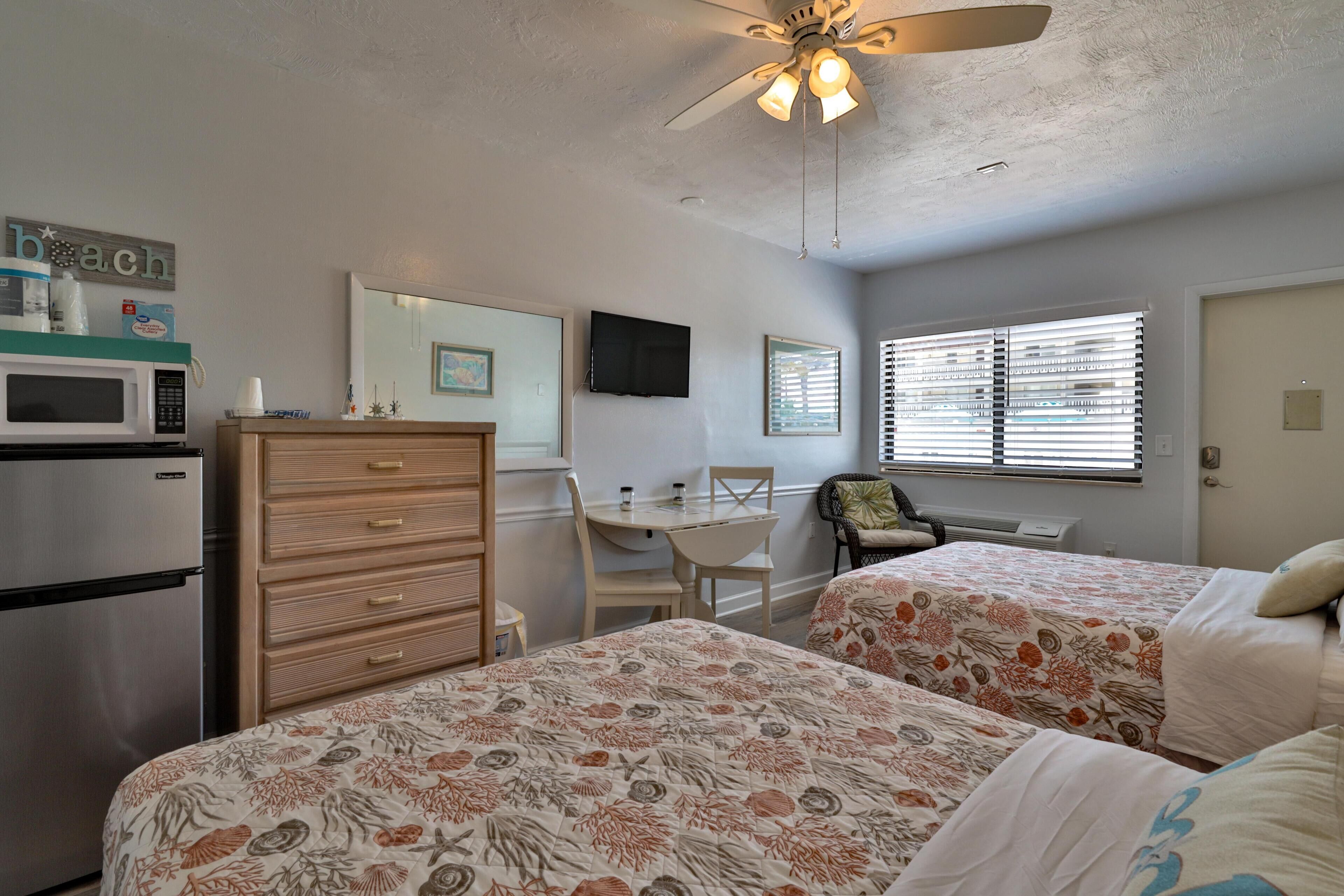 A bedroom with two beds, a desk, a chair, a microwave, a refrigerator, and a ceiling fan.
