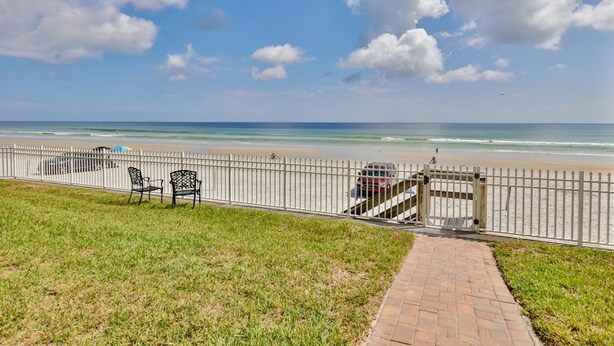 A beach view with a white fence, a paved walkway, and two metal chairs.