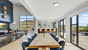 A spacious dining area with a long wooden table and blue chairs, large windows offering a view of the outdoors, and modern decor including pendant lights and wall-mounted shelves. - Epic 7BR Escape w/ Pickleball, Pool, Spa & Unmatched Mountain Views (Paradise Valley)