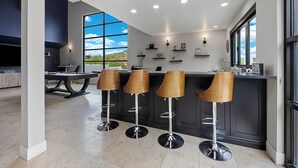 A modern kitchen with a bar area, featuring four wooden bar stools, a dining table, and a large window offering a view of the outdoors. - Epic 7BR Escape w/ Pickleball, Pool, Spa & Unmatched Mountain Views (Paradise Valley)