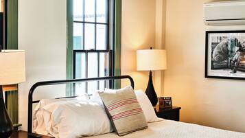A hotel room with a bed, a window, a wall-mounted air conditioner, and a framed picture.