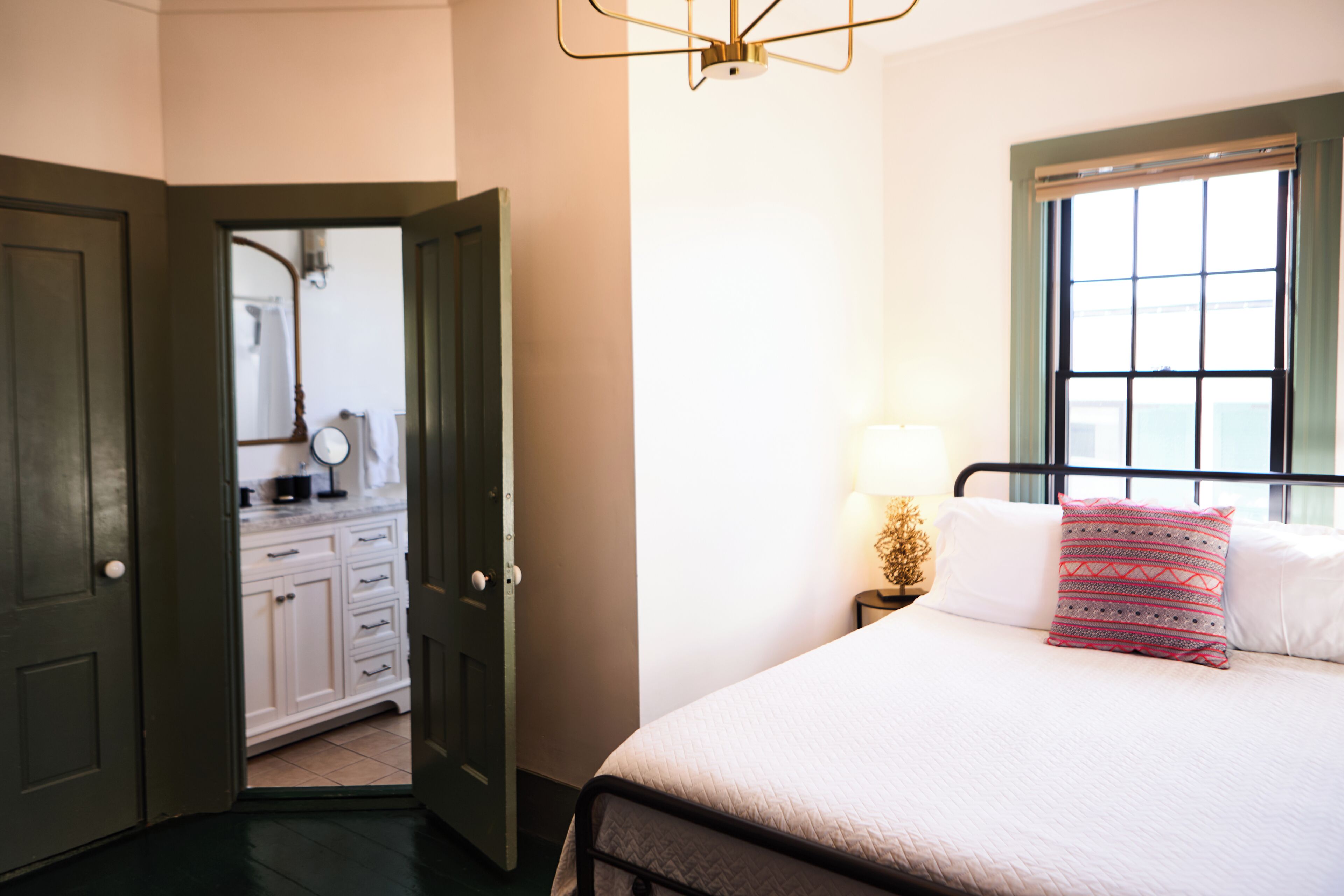 Un dormitorio con cama, mesita de noche, lámpara, ventana y un baño visible a través de una puerta abierta.