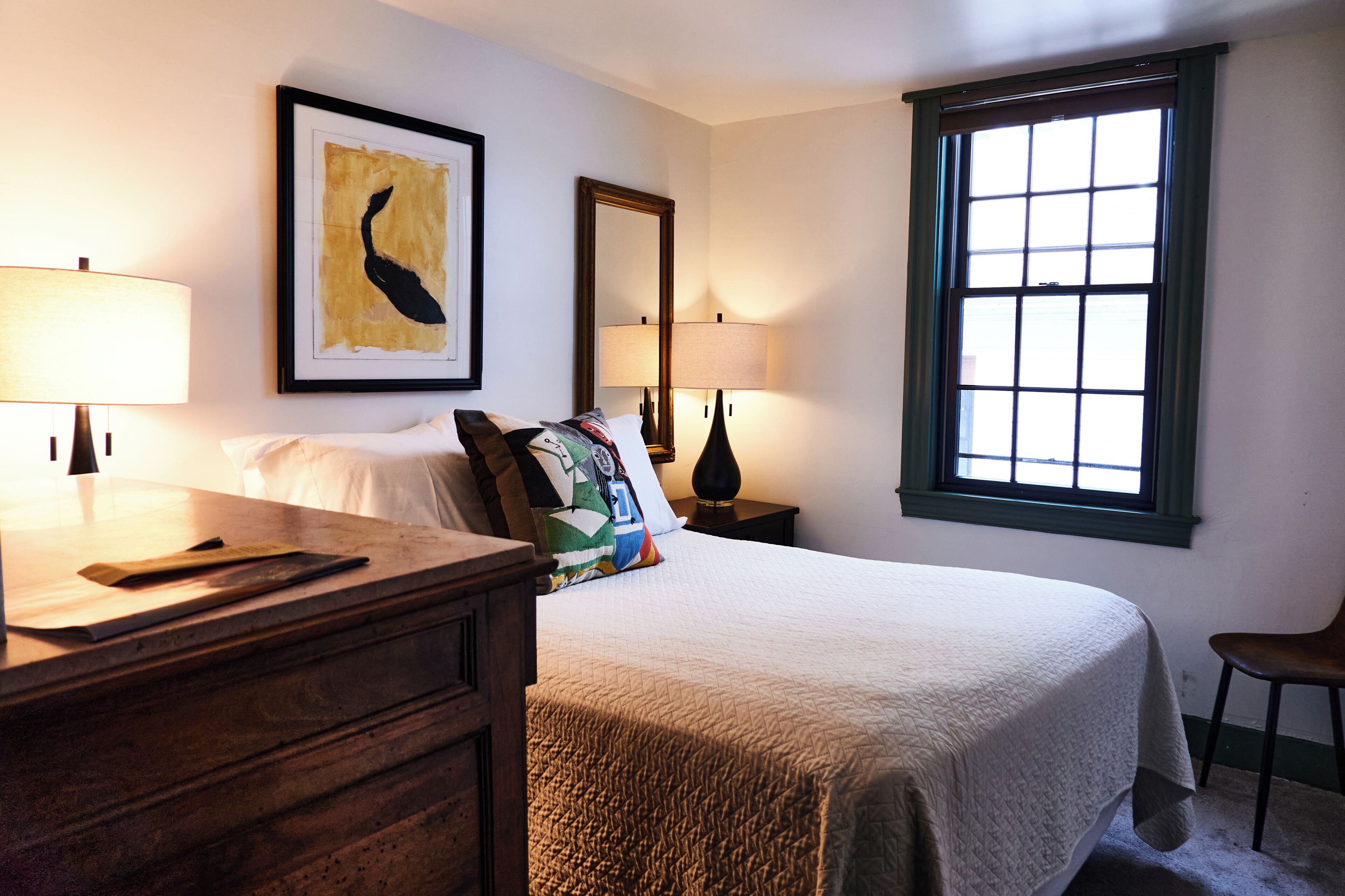A neatly arranged bedroom with a bed, bedside lamps, a dresser, a window with blinds, and a framed artwork on the wall.