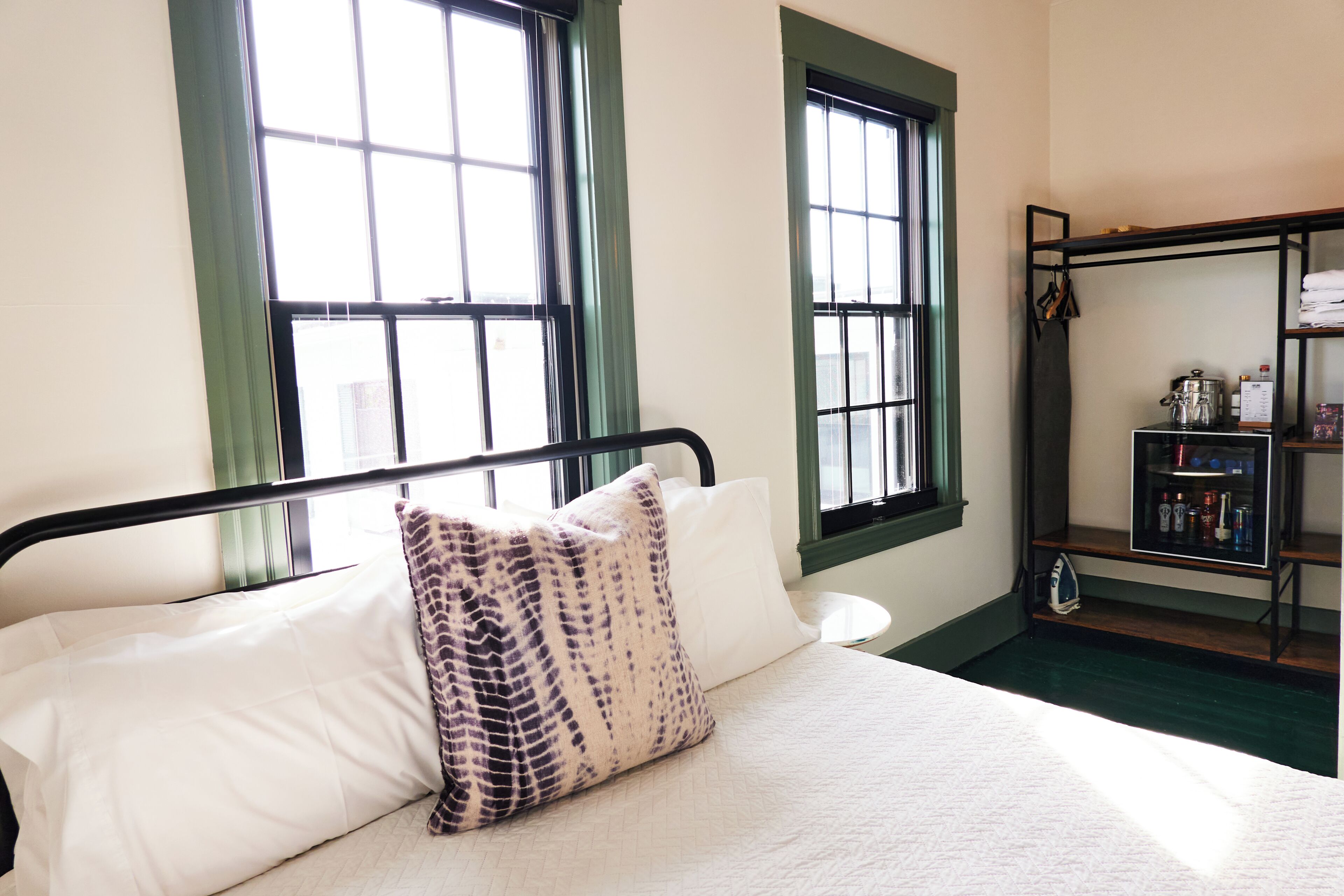 A hotel room with a bed, two windows with curtains, a small table, and a shelf with various items.