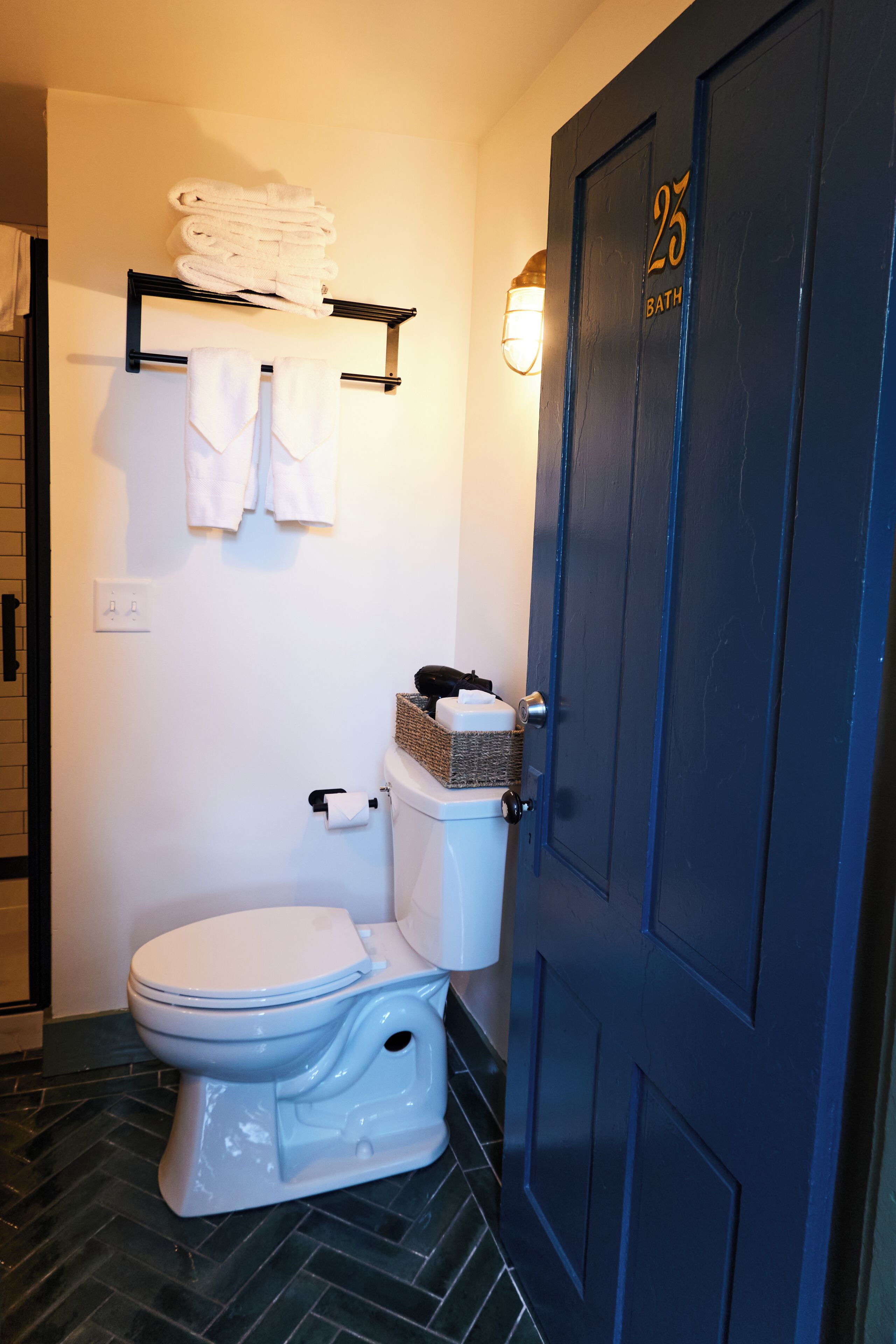 A bathroom with a toilet, a towel rack with folded towels, a door with a number, and a light fixture.