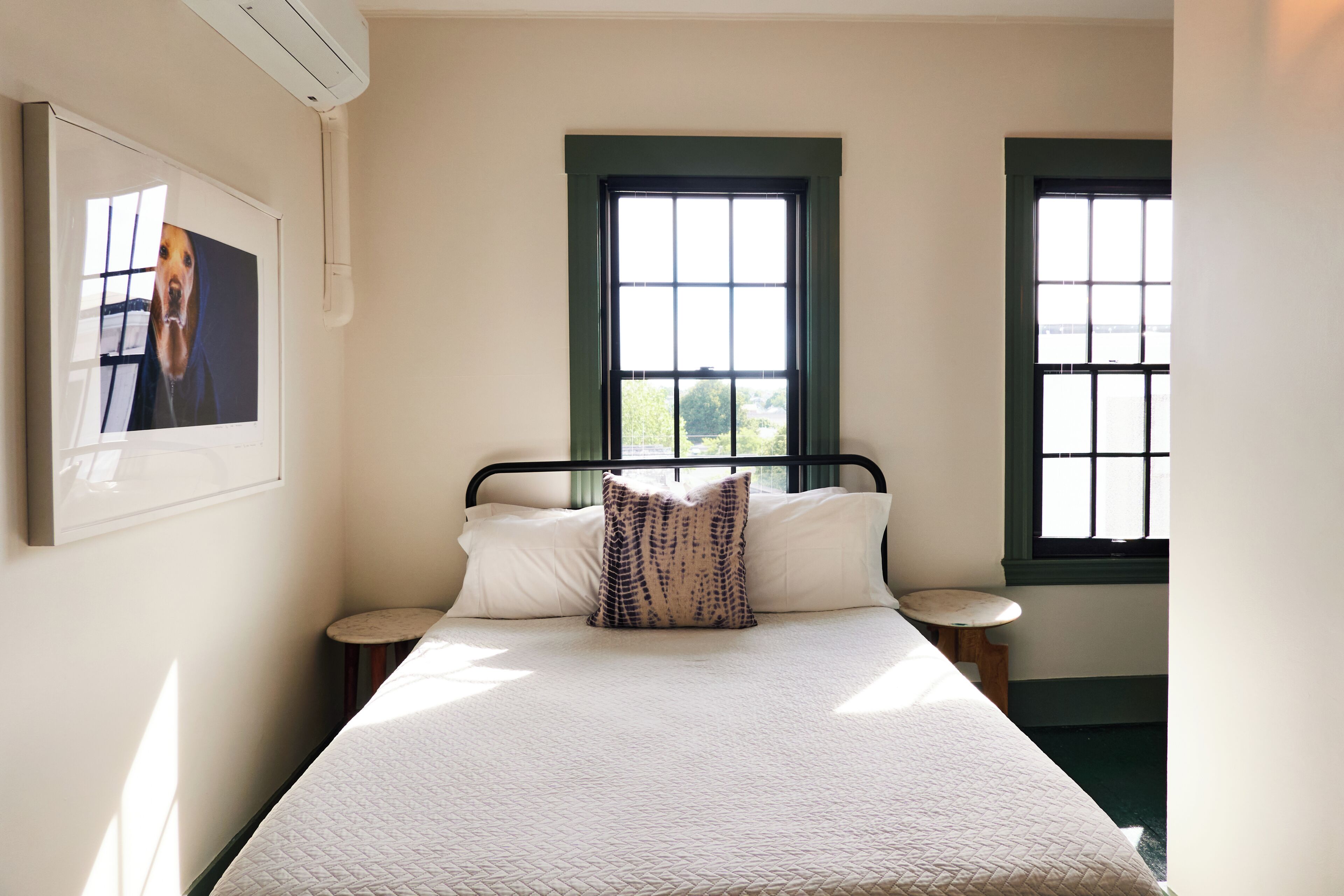A bedroom with a bed, two windows, a wall-mounted air conditioner, and a framed picture.