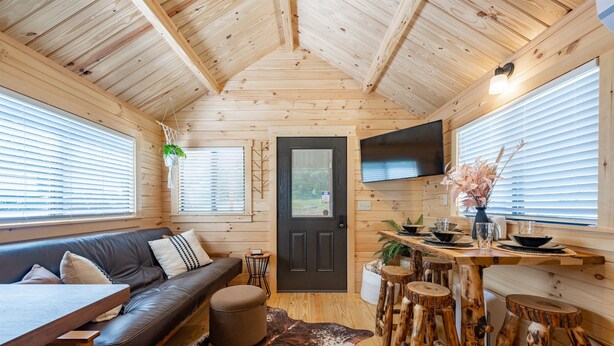L’intérieur chaleureux d’un chalet avec un plafond en bois, une porte noire, un canapé, une table à manger et un téléviseur fixé au mur.