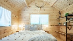 Une pièce chaleureuse, avec des murs en bois, comprenant un lit, des lampes de chevet, une fenêtre et une étagère ornée d’objets décoratifs.