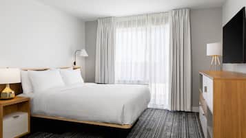 Quarto de hotel com cama, mesa de cabeceira com luminária, cômoda e televisão fixada na parede.