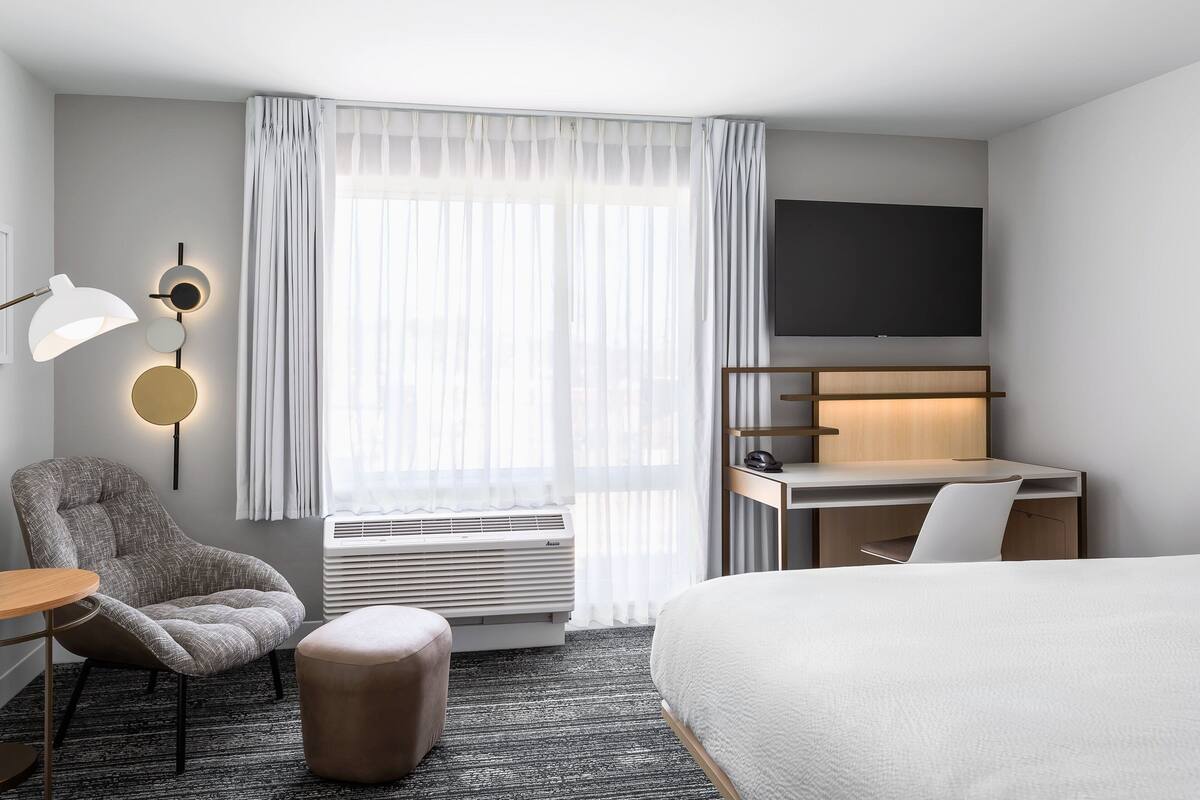 Een moderne hotelkamer met een bed, een bureau, een stoel, een lamp en een groot raam met dunne gordijnen.