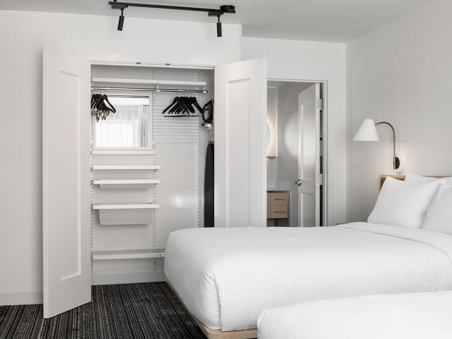 A hotel room with a bed, a closet with hanging clothes, a bedside table, and a lamp.