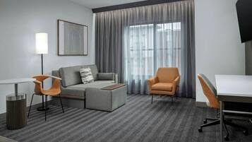 A modern hotel room with a grey sofa, two armchairs, a small table, a TV, and a large window with curtains.