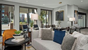 A modern living room with a sofa, a coffee table, and a dining area. There are large windows, a plant, and a lamp. - Courtyard by Marriott Bozeman (Bozeman)