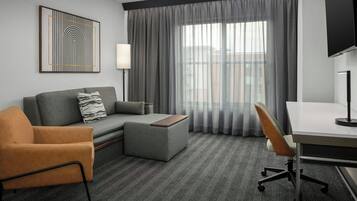 Una habitación de hotel con un sofá gris, un sillón naranja, un otoman gris, un escritorio con una silla, un televisor montado en la pared y un ventanal con cortinas translúcidas.