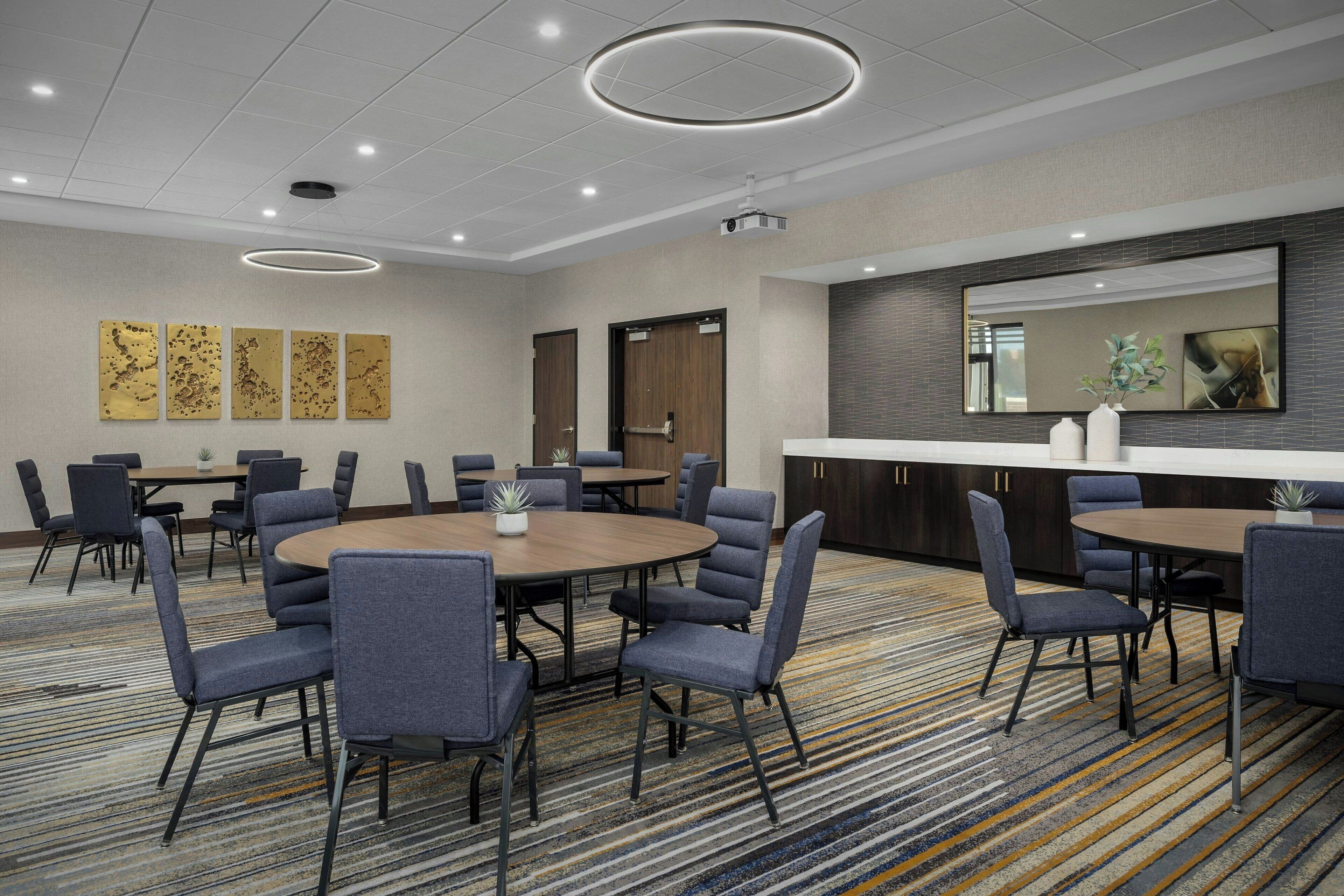 A conference room with round tables, chairs, a projector, and a large mirror on the wall.