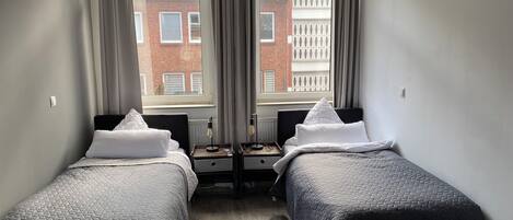 Une chambre d’hôtel avec deux lits, une fenêtre avec des rideaux, un radiateur et une vue sur un immeuble en briques.