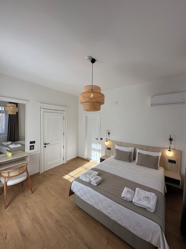 A modern hotel room with a large bed, a desk, and a chair. There is a door, a wall-mounted air conditioner, and a hanging light fixture. - Nueva Cunda (Ayvalik)