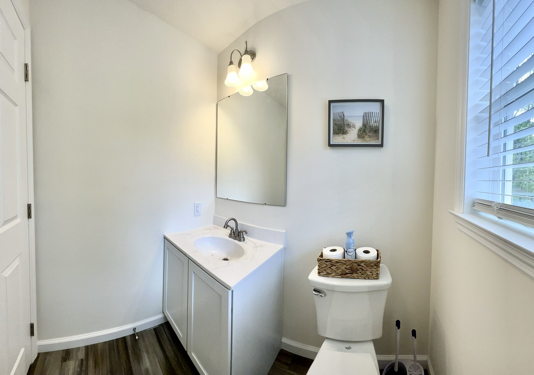 Ein Badezimmer mit weißem Waschbecken, einem Spiegel und einer Toilette. An der Wand hängt ein Bild und es gibt ein Fenster mit Jalousien.
