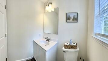 Ein Badezimmer mit weißem Waschbecken, einem Spiegel und einer Toilette. An der Wand hängt ein Bild und es gibt ein Fenster mit Jalousien.